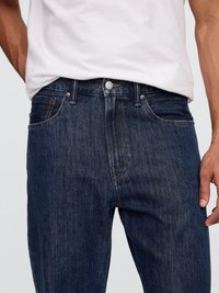 Dunkelblaue Jeans aus Denim mit einem klassischen Fünf-Taschen-Design, Knopfverschluss und subtiler Verwaschung. Getragen mit einem schlichten weißen T-Shirt.