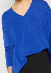 Femme portant un pull col en V bleu cobalt ample légèrement rentré dans un pantalon noir taille haute.