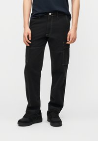 Άτομο που φοράει μαύρο cargo παντελόνι με πλαϊνές τσέπες και μαύρα αθλητικά παπούτσια, στέκεται μπροστά σε ένα απλό λευκό φόντο.