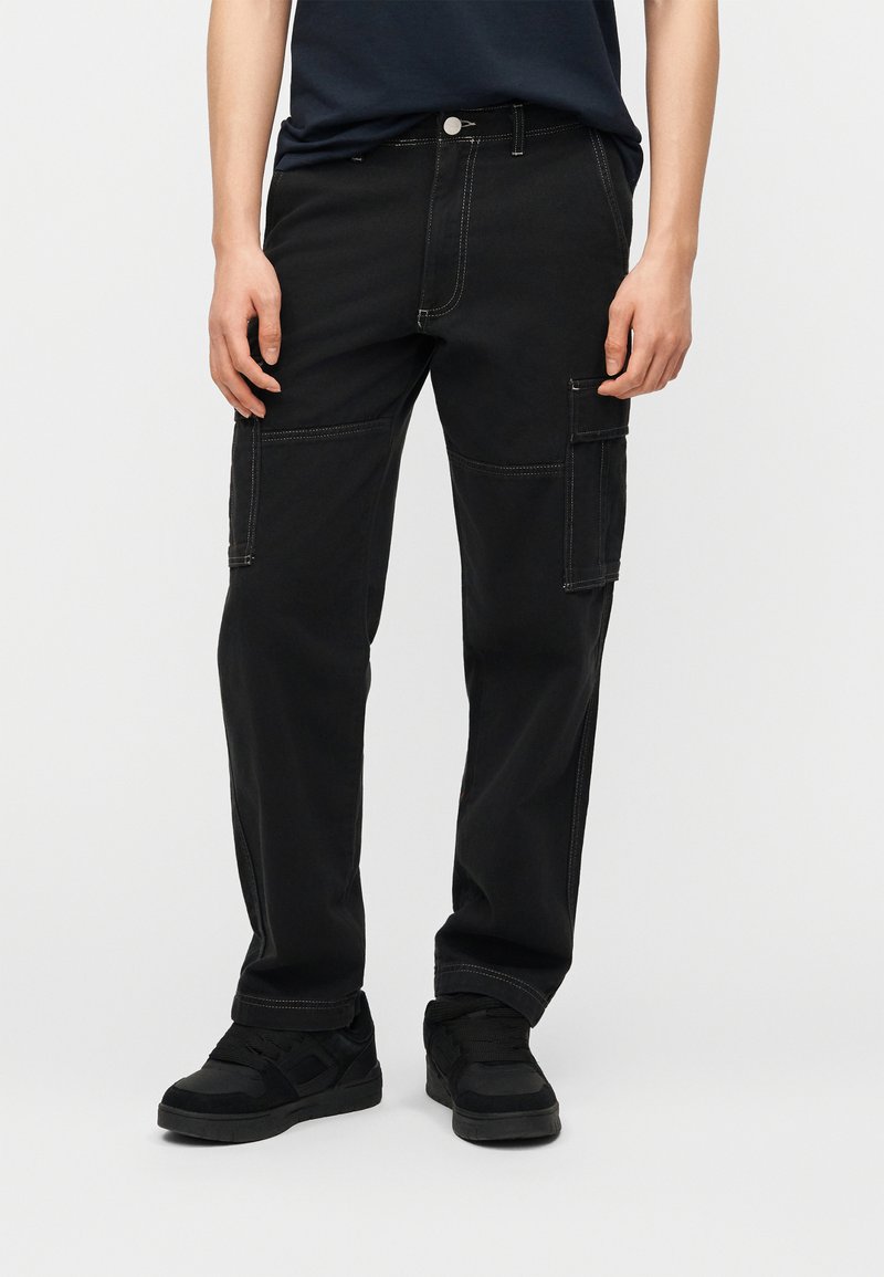 Άτομο που φοράει μαύρο cargo παντελόνι με πλαϊνές τσέπες και μαύρα αθλητικά παπούτσια, στέκεται μπροστά σε ένα απλό λευκό φόντο.