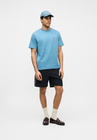 Homme portant une casquette et un t-shirt bleu clair, un short noir, des chaussettes crème et des mocassins marron foncé, debout devant un fond blanc uni.