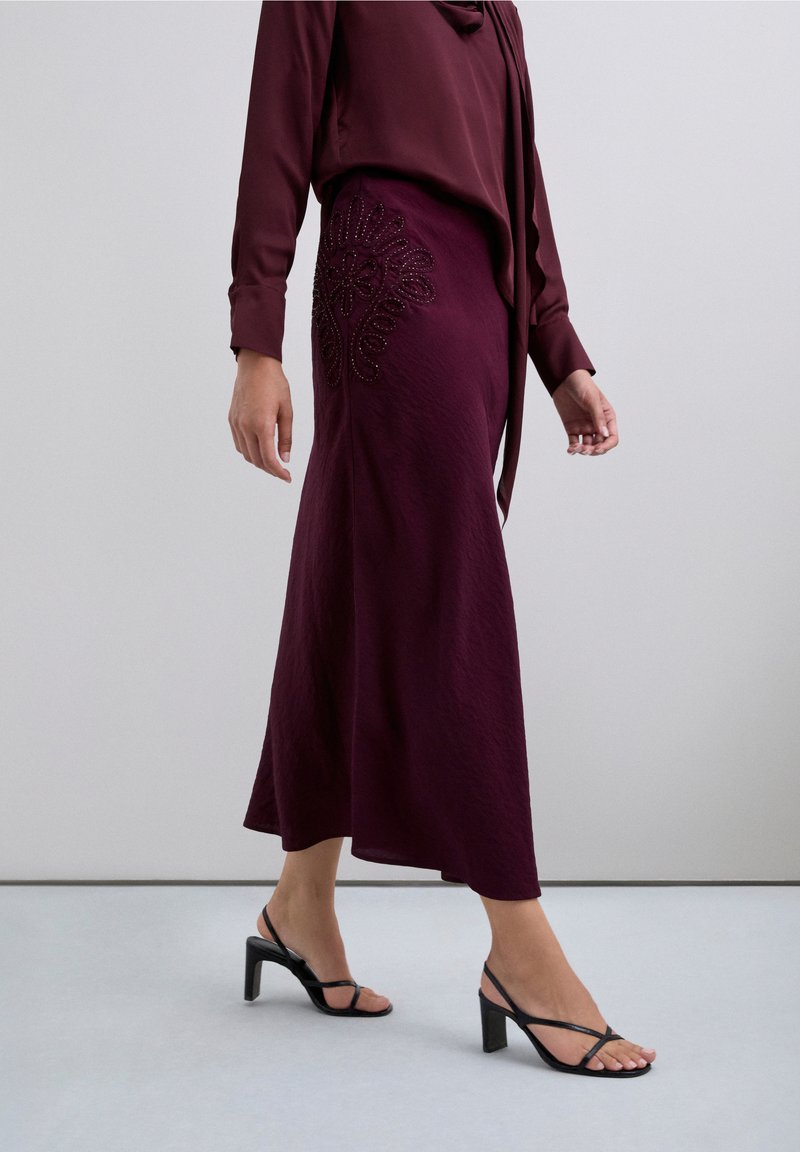 Falda color vino con tela texturizada y detalles de cuentas intrincadas en el costado, combinada con tacones negros con tiras y una blusa a juego.