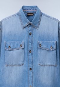 Lyseblå denimskjorte med spidse krave, to brystlommer, knaplukning foran og mørke knapdetaljer. Glat stofstruktur.