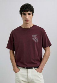 Camiseta de algodón burdeos de manga corta, con un gráfico blanco de dados y el texto "SCALPERS GAME" en la parte izquierda del pecho.