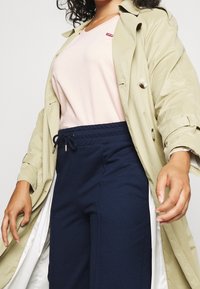 Trench coat beige, t-shirt rosa chiaro con un piccolo logo rosso e pantaloni jogger blu navy con cintura elastica.