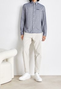 Šedá košile s knoflíky a malým logem "BOSS", kombinovaná s krémovými kalhotami a bílými teniskami na neutrálním pozadí.