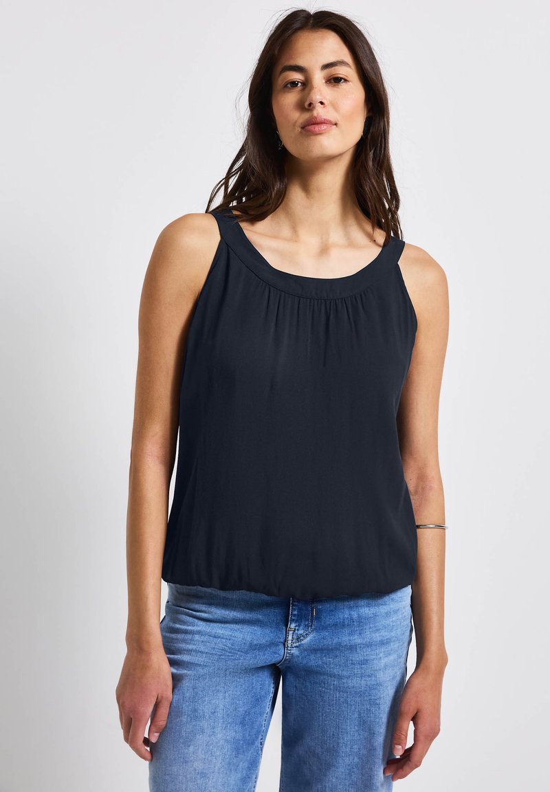 Blusa negra sin mangas con escote redondeado, textura fruncida en la parte delantera y corte holgado en la cintura, combinada con jeans de mezclilla.