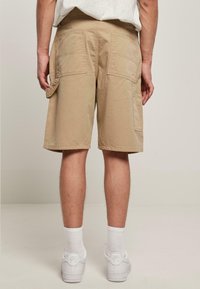 Beige cargo šortky s dvěma velkými zadními kapsami, volným střihem, zesíleným šitím a bočními smyčkami na nářadí nebo doplňky.