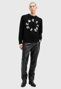 Sort strikket sweater med hvid cirkulær grafisk tekst, parret med sorte læderbukser og sorte ankelstøvler med en sølv kæde detalje.