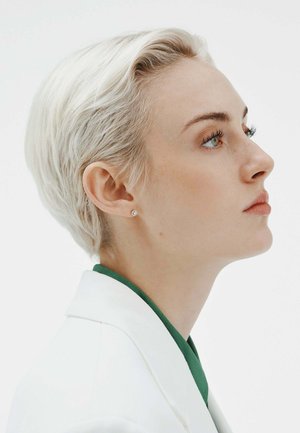 Blazer blanco corto, camisa verde debajo, cabello rubio claro corto y pendientes de tuerca. Fondo neutro, vista de perfil del sujeto.