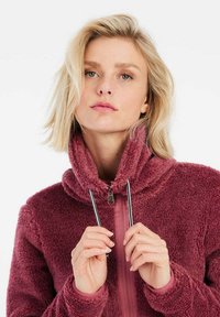 Giacca in fleece di un rosa intenso con un colletto alto, chiusura a zip e lacci. Superficie morbida e strutturata con accenti in tessuto contrastante.