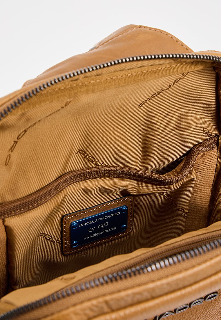 Interno di una borsa in pelle marrone con rivestimento in tessuto tan, dotata di un'etichetta metallica con la scritta "PIQUADRO QV 0378" e dettagli di zip liscia.