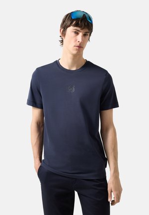 Młody mężczyzna ubrany w granatowy t-shirt z małym logo na piersi i niebieskie lustrzane okulary przeciwsłoneczne spoczywające na głowie.