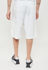 Pantaloni corti bianchi in cotone con vestibilità rilassata, dotati di una vita elasticizzata e dettaglio in rilievo del logo sul lato sinistro. Abbinati a sneakers nere.