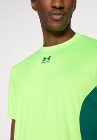 Αθλητική μπλούζ α με κοντά μανίκια από φθορίζον πράσινο πολυεστέρα, με σκουροπράσινη λεπτομέρεια στο πλάι και το λογότυπο της Under Armour στο στήθος.