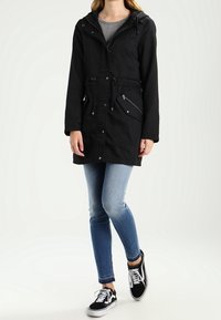 Svart vattentät parka med huva, dragsko i midjan, fickor med dragkedja och silverdetaljer. Bärs över en ljusgrå topp och jeans.