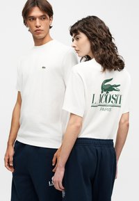 Νεαρός άνδρας και γυναίκα που φορούν λευκά T-shirts Lacoste και μπλε σκούρες παντελόνες, η γυναίκα δείχνει το λογότυπο και το κείμενο Lacoste στην πλάτη του πουκάμισού της.