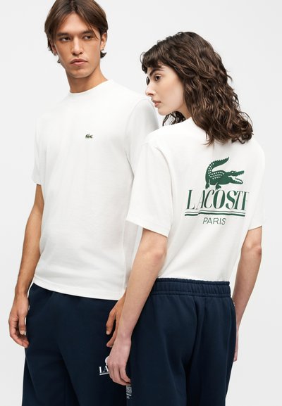 Млад мъж и жена, носещи бели тениски Lacoste и тъмносини панталони, жената показва логото и текста на Lacoste на гърба на тениската си.