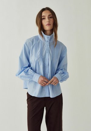 Kvinde med langt hår iført lyseblå højhalset bluse med folder og knapper, kombineret med mørke bukser, stående foran ensfarvet baggrund.