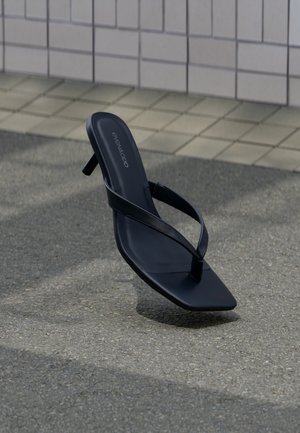 Μαύρο σανδάλι με τακούνι, λεπτά λουράκια και τετράγωνη μύτη, στέκεται όρθιο σε υφή πεζοδρομίου κοντά σε πλακιδωτό τοίχο.