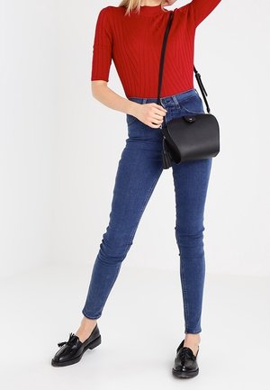 Nainen, joka pukeutunut punaiseen lyhythihaiseen neuleeseen, sinisiin kapeisiin farkkuihin, mustiin loafereihin ja mustaan olkalaukkuun, seisoo yhdellä kädellä ylös kohotettuna.