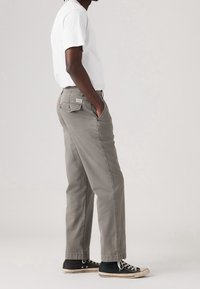 Man wearing white t-shirt, gray straight-leg pants, black high-top sneakers, standing with hands in pockets against plain background.