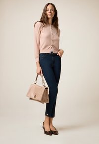 Cardigan rose clair avec des boutons dorés, jean en denim foncé, sac beige avec des détails en métal, et ballerines pointues marron sur un fond neutre.