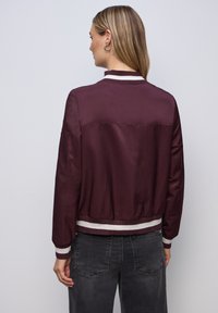 Femme aux longs cheveux portant une veste bomber bordeaux avec des rayures blanches sur le col, les poignets et la ceinture, assortie à un jean noir, vue de dos.