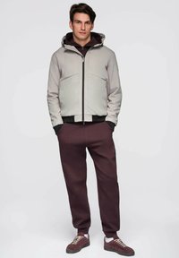 Jasnoszara kurtka z kapturem, czarnymi akcentami, zamek błyskawiczny i dwie przednie kieszenie; zestawiona z ciemnoczerwonymi spodniami dresowymi i brązowymi trampkami.