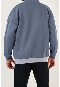 Sudadera de color azul claro con un cuello alto, puños y dobladillo acanalados. Presenta una textura suave y un corte holgado, y está diseñada sin logotipos visibles.