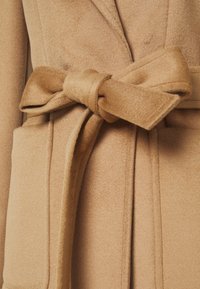 Kameliinivärinen pitkä takki pehmeällä tekstuurilla, jossa on solmittava vyö ja kaksi sivutaskua. Halkiokaulus ja siistit ompelusaumat.