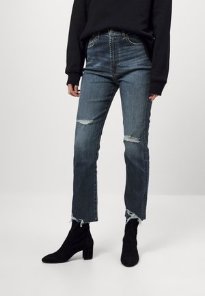 Jeans de tiro alto en azul oscuro con desgastes, dobladillo deshilachado y una textura destrozada, combinados con botas negras de tobillo.