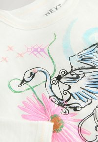 T-shirt en coton blanc arborant une illustration colorée d'un cygne, une fleur rose, des vignes vertes et des accents bleus avec des contours noirs. Étiquette suivante visible.