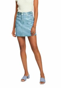 Femme portant une minijupe en denim bleu clair brillant, un pull sans manches blanc, des bagues et des sandales à talons carrés bleu clair.