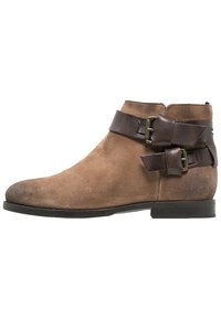 Stivaletto in suede marrone con punta rotonda, doppie cinghie in pelle marrone con fibbie metalliche e suola piatta con minimalissimo battistrada.