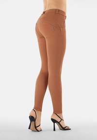 Leggings aderenti marroni con una texture liscia, caratterizzati da un design con cuciture curve e dettagli in metallo argentato sulle tasche posteriori. Abbinati a tacchi neri alti.