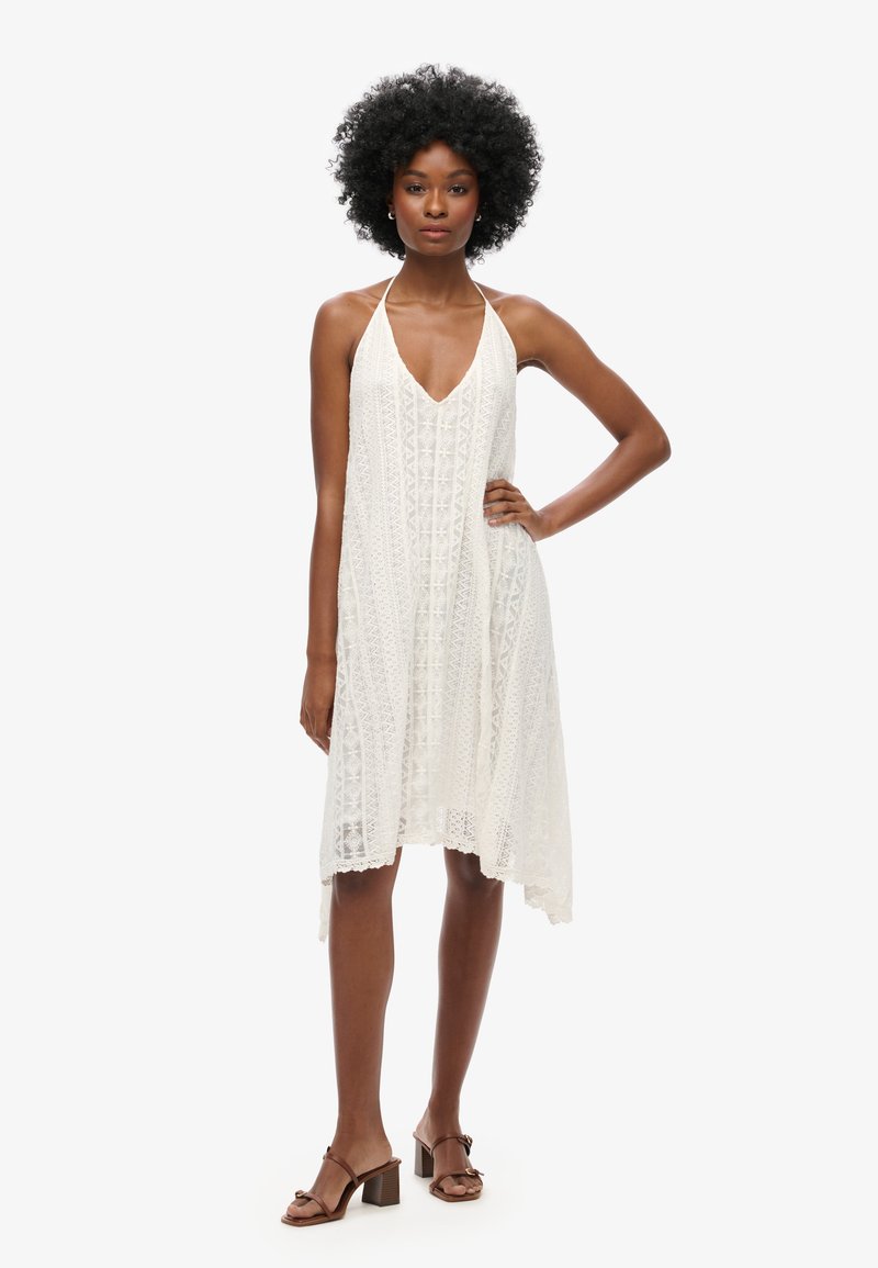 Weißes Spitzenkleid mit V-Ausschnitt, Trägern im Halter-Design und einer fließenden Silhouette. Verfügt über aufwendige Muster und einen Saum, der hinten tiefer ausfällt.