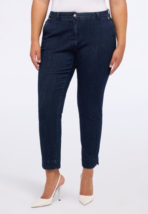 Frau in dunkelblauen Jeans und weißen, hochhackigen Slingback-Schuhen, steht vor einem schlichten weißen Hintergrund.