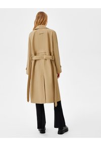 Manteau beige en tissu lisse, avec une ceinture, une fente au dos et un large col. Stylisé avec un pantalon noir et des chaussures épaisses.