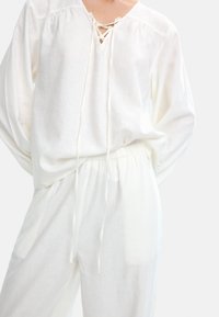 Biała, lekka bluzka z dekoltem w serek i sznurowanym detalem, zestawiona z luźnymi białymi spodniami z elastycznym pasem.
