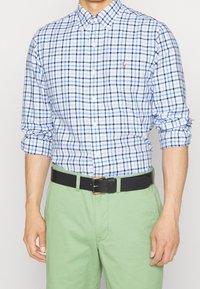 Knappad rutig skjorta i ljusblått och marinblått, med krage, långärmar uppkavlade, i kombination med gröna chinos och ett svart bälte.
