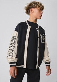 Chaqueta tipo varsity negra y crema con botones a presión, mangas en contraste, texto "BAKER" en la manga y detalles en los bolsillos. Textura de tela suave.