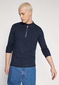 Pulover tricotat bleumarin cu guler cu fermoar, având mâneci lungi și un design ajustat. Textură netedă și stil minimalist.