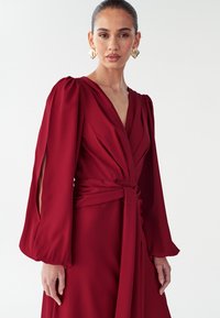 Punane wrap-kleit, mis on valmistatud siledast kangast, millel on pikad puhvis varrukad, sügav V-kaelus ning vöökoht nööriga, mis annab vormi järgiva silueti.