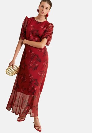 Maxime dress in red with floral pattern, sheer puff sleeves, and layered hem. Paired with a gold clutch and red heeled sandals.