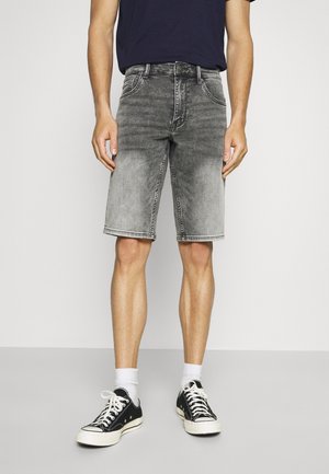 Homme portant un short en denim noir délavé, des chaussettes blanches mi-mollet et des baskets basses noires, sur un fond léger uni.