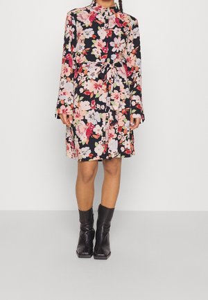 Femme portant une robe noire mi-longue avec un motif floral coloré, des manches longues, une ceinture à la taille et des bottes en cuir noires mi-mollet.