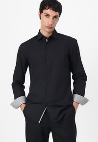 Black long-sleeve shirt with button closure, contrasting patterned cuff, and collar detail, made of smooth fabric. Tailored fit design.