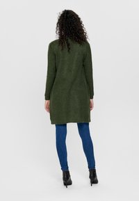Langer grüner Cardigan aus strukturiertem Strickmaterial, mit langen Ärmeln und lockerer Passform, getragen über blauen Skinny-Jeans und schwarzen Knöchelstiefeln.