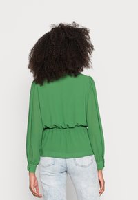 Grüne Bluse mit langen Ballonärmeln, tailliertem Bund und glattem, leichtem Stoff. Getragen mit hellblauen Jeans. Rückansicht.